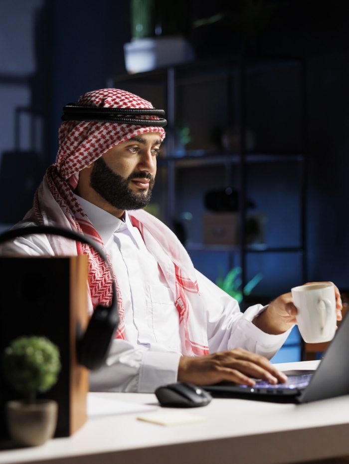 Arab guy holding a cup and using laptop Side-view of a Muslim guy holding a cup of coffee and surfing the net on his digital laptop. Image showcasing a male Arab person engrossed in his personal computer while drinking tea.